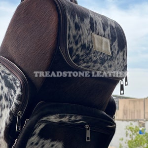 Pu&ograve; includere: Uno zaino in pelle di mucca marrone e bianco con una tasca con cerniera e il testo "Treadstone Leather" sul davanti.