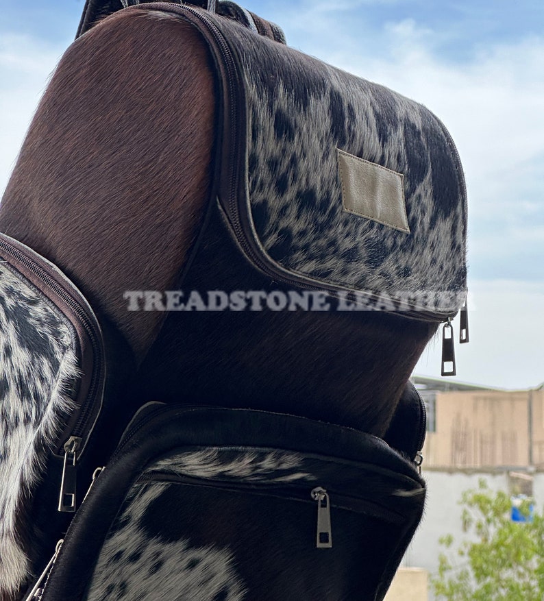 Pu&ograve; includere: Uno zaino in pelle di mucca marrone e bianco con una tasca con cerniera e il testo "Treadstone Leather" sul davanti.