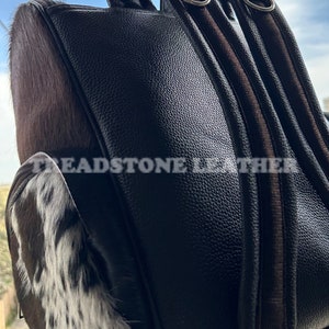 Pu&ograve; includere: Uno zaino in pelle marrone con una toppa in pelle di mucca sul davanti. Lo zaino ha due spallacci regolabili e una tasca con cerniera sul davanti. Il testo "Treadstone Leather" &egrave; visibile sul lato dello zaino.