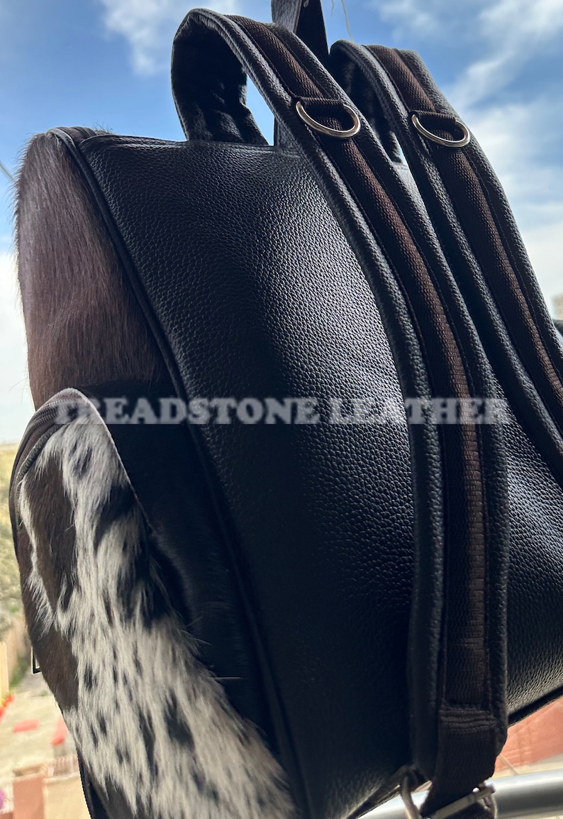 Pu&ograve; includere: Uno zaino in pelle marrone con una toppa in pelle di mucca sul davanti. Lo zaino ha due spallacci regolabili e una tasca con cerniera sul davanti. Il testo "Treadstone Leather" &egrave; visibile sul lato dello zaino.