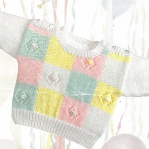 May include: A white knitted baby sweater with a colorful checkered pattern in pink, yellow, and green. The sweater has a round neckline and long sleeves. There are small white buttons on the shoulders.