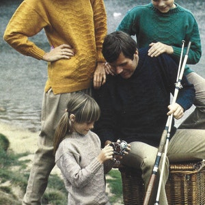 May include: A family of four wearing sweaters, with a child holding a fishing reel. The adults are standing, while the child is sitting on a wicker basket. The family is outdoors, with a body of water in the background.