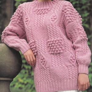 以下が含まれることがあります： ケーブル編みのパターンとボブルが付いたピンクのニットセーター。セーターは丸首と長袖です。
