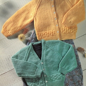 May include: Two knitted cardigans, one yellow and one green, with button closures. Both cardigans have a ribbed texture and are laid out on a patterned fabric.