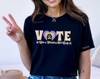 May include: Black t-shirt with the text "VOTE Like a Childless Cat Lady" in a retro font. The word "VOTE" has a rainbow heart with a black paw print in the center.