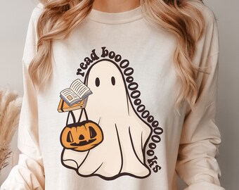 May include: Cream-colored long-sleeve shirt featuring a cartoon ghost holding a book and a jack-o'-lantern. The ghost has the words "read boooooooks" around it. The shirt is a Halloween-themed design.