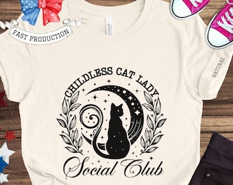 May include: A white t-shirt with a black graphic design featuring a cat sitting on a crescent moon with stars and the text "Childless Cat Lady Social Club".