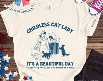 May include: A white t-shirt with a blue graphic of a woman with a basket of kittens. The text "Childless Cat Lady" is above the graphic and "It's a beautiful day to stop the patriarchy one kitten at a time" is below the graphic.