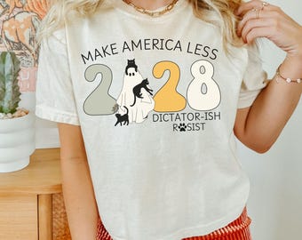 May include: Cream-colored t-shirt with the text "MAKE AMERICA LESS 2028 DICTATOR-ISH RESIST". The design features a ghost with black cats and the numbers 2028 in gray, yellow, and white.