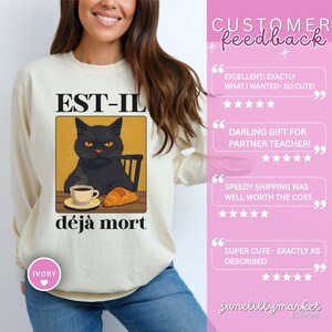 May include: Ivory-colored sweatshirt featuring a graphic of a black cat with orange eyes, a cup of coffee, and a croissant. The text "EST-IL d&eacute;j&agrave; mort" is above and below the cat illustration. The sweatshirt has long sleeves.