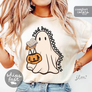 May include: A cream-colored short-sleeve t-shirt featuring a graphic of a white ghost holding an open book and an orange pumpkin bucket. The text "read BOOOOOOOOOKS" is printed in black, curving around the ghost. The shirt has a crew neckline.