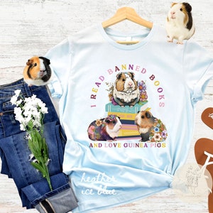 May include: Light blue t-shirt with the text "I read banned books and love guinea pigs" in a circle around a stack of books with three guinea pigs sitting on top. The guinea pigs are wearing flowers and the books are decorated with flowers.