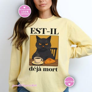 May include: A light yellow long-sleeve shirt featuring a graphic of a black cat with orange eyes, a cup of coffee, and a croissant. The text "EST-IL d&eacute;j&agrave; mort" is above and below the cat illustration. The shirt is made of 100% cotton.