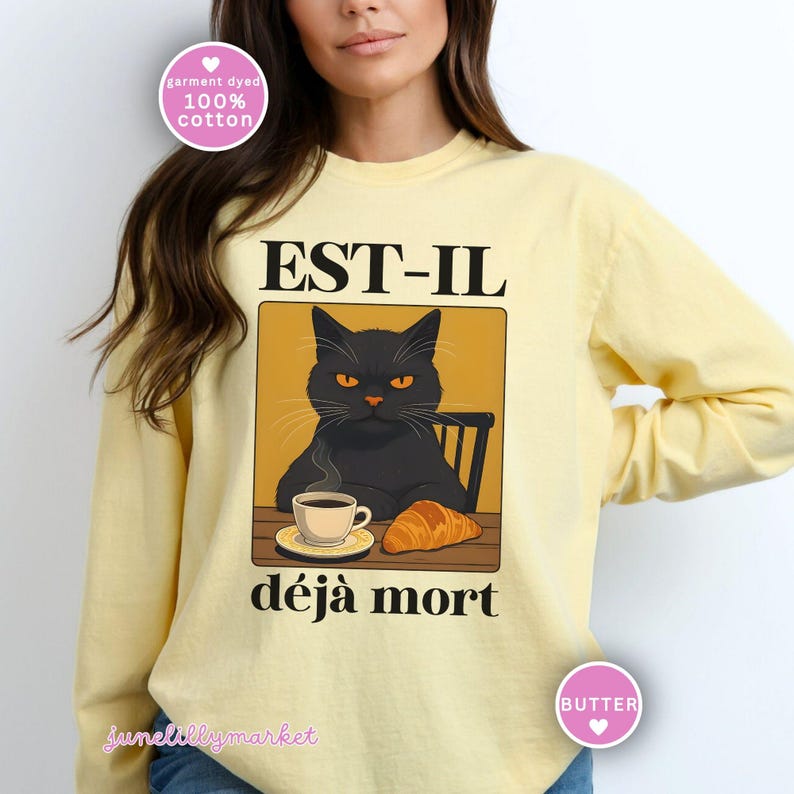 May include: A light yellow long-sleeve shirt featuring a graphic of a black cat with orange eyes, a cup of coffee, and a croissant. The text "EST-IL d&eacute;j&agrave; mort" is above and below the cat illustration. The shirt is made of 100% cotton.