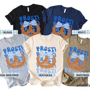 May include: A collection of t-shirts in various colors, including black, navy, and heather olive. Each shirt features the word "PROST!" in blue, along with a pretzel design and a blue and white bow. The shirts are from junelillymarket.