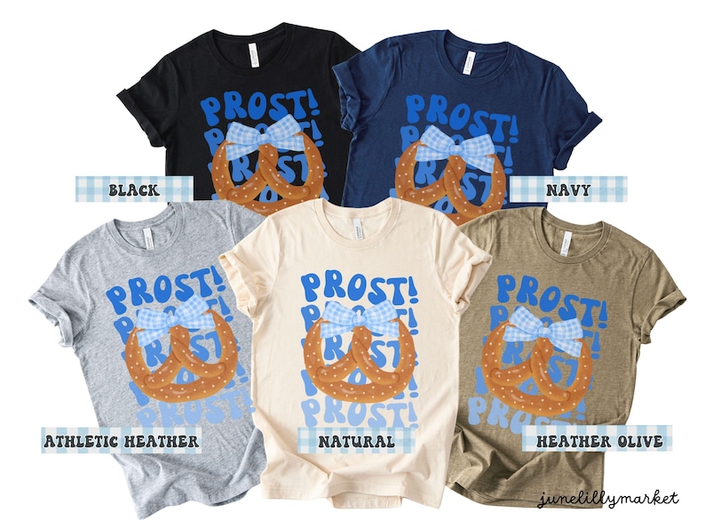May include: A collection of t-shirts in various colors, including black, navy, and heather olive. Each shirt features the word "PROST!" in blue, along with a pretzel design and a blue and white bow. The shirts are from junelillymarket.