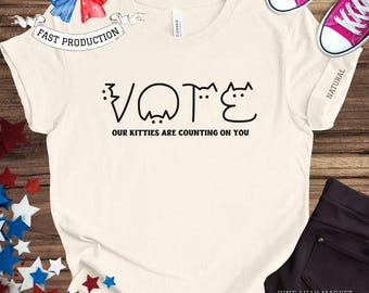 May include: A white t-shirt with a black graphic of the word "VOTE" spelled out using cat silhouettes. The text "Our kitties are counting on you" is printed below the graphic.