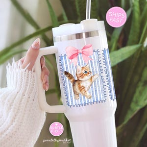 May include: White insulated tumbler with a handle, featuring a watercolor illustration of a playful orange tabby kitten with a pink bow. The design includes blue stripes and decorative swirls. The tumbler has a white lid and a straw.