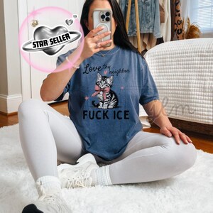 May include: A blue t-shirt with the text "Love thy neighbor" above a graphic of a kitten wearing a pink bow. Below the kitten is the text "FUCK ICE". The shirt is worn with white leggings and sneakers.
