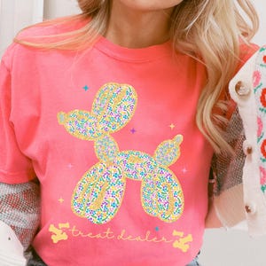 May include: A coral pink t-shirt featuring a balloon dog design with a floral pattern in yellow, blue, and purple. The text "treat dealer" is below the dog. The shirt has a crew neck and short sleeves.