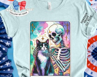 Pode incluir: Uma camiseta azul claro com um gráfico de um esqueleto segurando um gato preto e branco. O gráfico é cercado por um fundo colorido e estrelado. O texto "Childless Cat Lady" é impresso abaixo do gráfico.