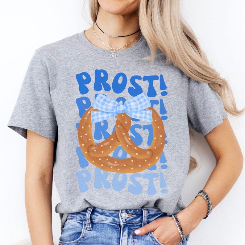 May include: A heather gray t-shirt featuring a pretzel design with a blue and white gingham bow. The word "PROST!" is printed in blue above and below the pretzel graphic. The shirt is made of a soft material.