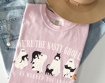 May include: A light pink t-shirt with the text "WE'RE THE NASTY GHOULS" and "45 WARNED YOU ABOUT". The graphic features white ghosts with black cats. A tan hat and blue jeans are in the background.