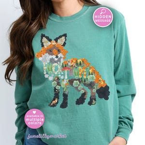 May include: Teal long-sleeve sweatshirt featuring a detailed fox design composed of flowers, foliage, and woodland elements. The fox illustration includes orange, white, and black accents. The text "available in multiple colors" is also visible.