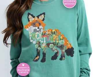 May include: Teal long-sleeve sweatshirt featuring a detailed fox design composed of flowers, foliage, and woodland elements. The fox illustration includes orange, white, and black accents. The text "available in multiple colors" is also visible.