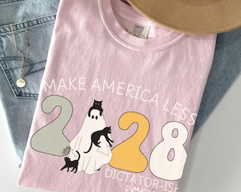 May include: A light pink t-shirt with the text "MAKE AMERICA LESS 2020 DICTATOR-ISH RESIST". The graphic includes a ghost with black cats and the numbers 2020 in gray and yellow. A tan hat and blue jeans are in the background.