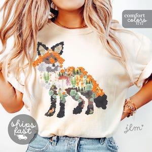May include: A cream-colored t-shirt featuring a graphic of a fox silhouette composed of orange flowers, green succulents, ferns, pine trees, and mushrooms. The text 'comfort colors 1717' appears in the top right corner, and 'ships fast' is in the bottom left.