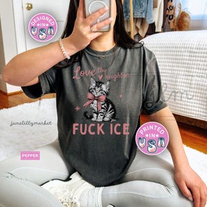 May include: Dark gray t-shirt with a graphic of a kitten wearing a pink bow and the text "FUCK ICE". The shirt also features the text "Love thy neighbor". Designed and printed in the USA.