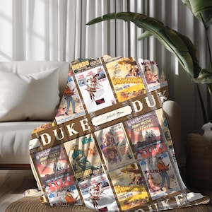 May include: A brown and tan throw blanket with a collage of vintage movie posters featuring John Wayne. The posters include "Stagecoach", "The Searchers", "Hondo", and "McLintock!". The blanket has a brown and white plaid pattern and the text "Duke" is repeated throughout.
