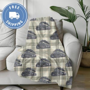May include: A soft blanket draped over a sofa, featuring a repeating pattern of vintage steam trains on a plaid background. The trains are depicted in shades of gray and black, with puffs of smoke. The blanket adds a cozy touch to the living space.
