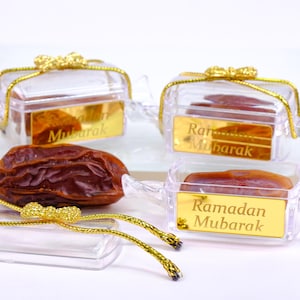 Peut inclure: Quatre boîtes à bonbons en plastique transparent avec des nœuds en ruban doré et des inscriptions dorées. Les boîtes sont en forme de bonbon et portent les mots "Ramadan Mubarak". Il y a une datte séchée devant les boîtes.