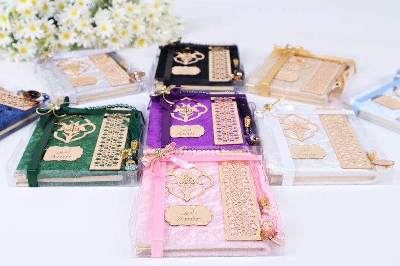 Op de afbeelding: Assortiment gebedsets in verschillende kleuren, elk met een fluwelen hoes, een houten bladwijzer en gebedskralen. De sets zijn verpakt in doorzichtige plastic dozen met een lint en een naamsticker met de tekst "Amir".