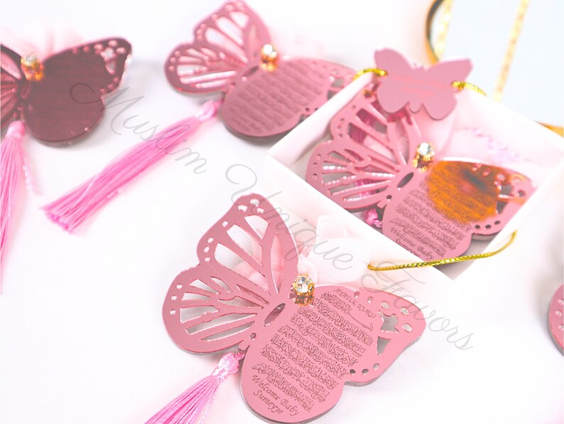K&ouml;nnte beinhalten: Rosa Schmetterlingsf&ouml;rmige Ornamente mit einem goldenen Quasten und einem Diamanten-f&ouml;rmigen Schmuckst&uuml;ck. Die Ornamente zeigen arabische Kalligraphie und den Text "Willkommen Baby Sumeyye".