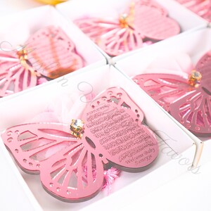 K&ouml;nnte beinhalten: Rosa Schmetterlingsf&ouml;rmige Ornamente mit einem dekorativen Design und einem kleinen Strassstein. Die Ornamente befinden sich in einzelnen Schachteln mit einem rosa Band und der Text "Welcome Baby Sameeye" ist auf das Ornament graviert.