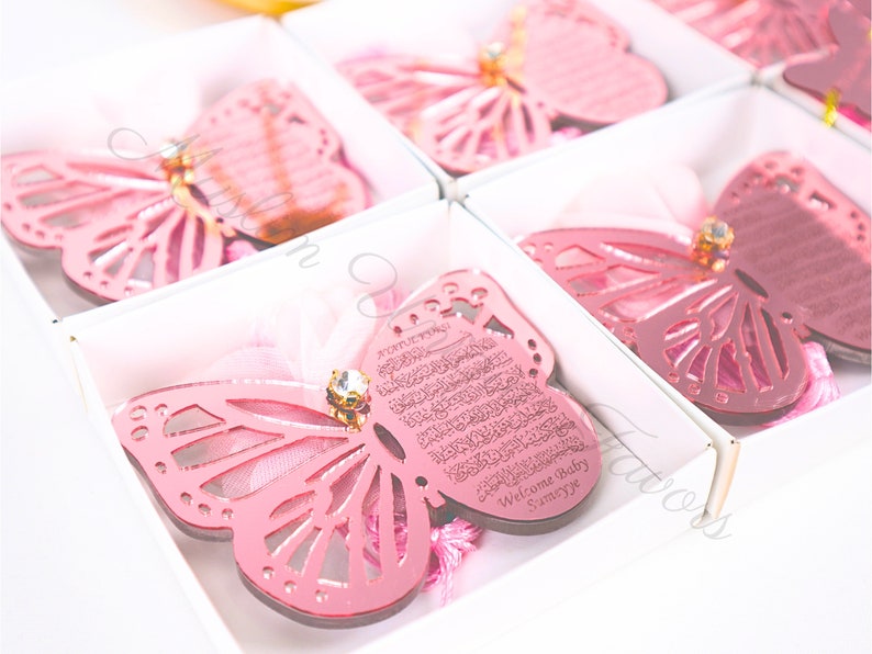K&ouml;nnte beinhalten: Rosa Schmetterlingsf&ouml;rmige Ornamente mit einem dekorativen Design und einem kleinen Strassstein. Die Ornamente befinden sich in einzelnen Schachteln mit einem rosa Band und der Text "Welcome Baby Sameeye" ist auf das Ornament graviert.