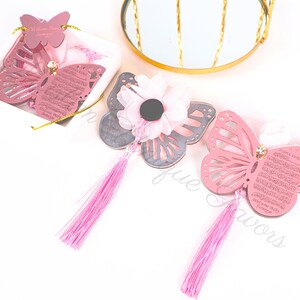 K&ouml;nnte beinhalten: Drei rosafarbene Schmetterlings-Anh&auml;nger mit je einem Quasten-Anh&auml;nger. Die Schmetterlinge haben ein ausgeschnittenes Design und sind mit arabischer Kalligraphie verziert. Ein Schmetterling befindet sich in einer wei&szlig;en Schachtel mit einem rosa Band.