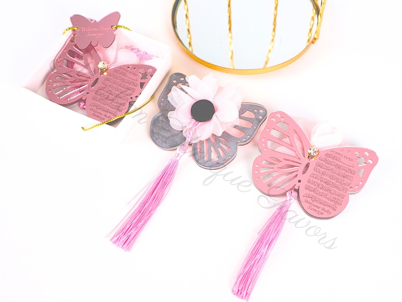 K&ouml;nnte beinhalten: Drei rosafarbene Schmetterlings-Anh&auml;nger mit je einem Quasten-Anh&auml;nger. Die Schmetterlinge haben ein ausgeschnittenes Design und sind mit arabischer Kalligraphie verziert. Ein Schmetterling befindet sich in einer wei&szlig;en Schachtel mit einem rosa Band.
