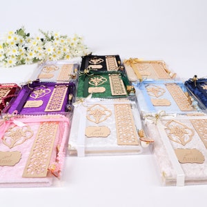 Op de afbeelding: Een assortiment gebedskleden in verschillende kleuren, waaronder roze, wit, blauw, groen, paars en rood. Elk gebedskleed is verpakt in een doorzichtige plastic zak met een houten bladwijzer, een kwast en een klein houten bordje met de tekst "Amir" erop.