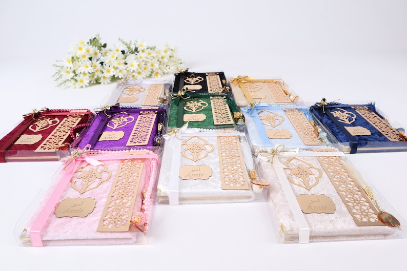 Op de afbeelding: Een assortiment gebedskleden in verschillende kleuren, waaronder roze, wit, blauw, groen, paars en rood. Elk gebedskleed is verpakt in een doorzichtige plastic zak met een houten bladwijzer, een kwast en een klein houten bordje met de tekst "Amir" erop.