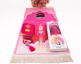 Peut inclure: Tapis de prière rose avec un motif de fusée, un livre de prière rose avec le nom "Zainab" dessus et des chapelets de prière roses dans une boîte blanche avec le nom "Zainab" dessus.
