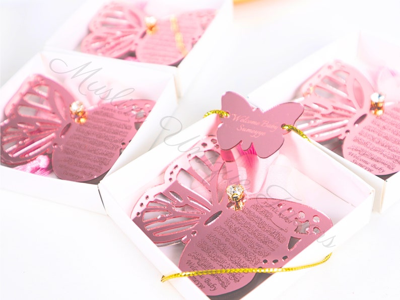 K&ouml;nnte beinhalten: Rosa Schmetterlingsf&ouml;rmige Geschenkboxen mit einem goldenen Band und einem kleinen diamantf&ouml;rmigen Strassstein. Die Boxen enthalten einen rosa Schmetterling mit der Aufschrift "Willkommen Baby" und eine rosa Karte mit arabischer Kalligraphie.