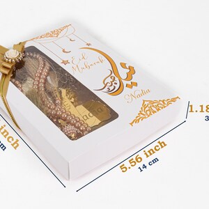 以下が含まれることがあります： 金色のリボンと透明な窓が付いた白いギフトボックス。窓からピンク色の数珠のセットが見えます。ボックスは金色の書道と「Eid Mubarak」と「Nadia」の文字で飾られています。ボックスのサイズは18cm x 14cm x 3cmです。
