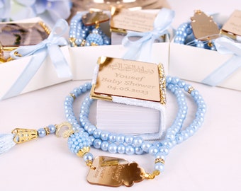 May include: A blue and gold baby shower favor with a personalized inscription that reads "Yousef Baby Shower 04.06.2023". The favor includes a small book with a white cover and a string of blue beads.