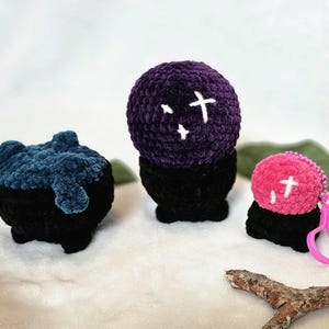 May include: Three crocheted cauldron-shaped plush toys with different colored tops. The first is blue, the second is purple with white crosses, and the third is pink with a white cross. The pink cauldron has a pink plastic keychain attached.
