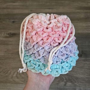 May include: A handmade crochet drawstring bag in a gradient of pastel colors: pink, purple, and teal. The bag has a textured, scaly pattern and a cream-colored drawstring closure. The bag is held in a hand.