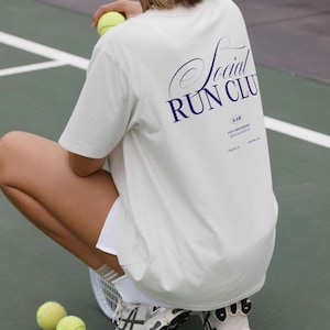 May include: Cream-colored t-shirt with the text "Social Run Club" in navy blue script. The shirt also features additional text and is paired with white shorts and sneakers. Several tennis balls are scattered on the green court.
